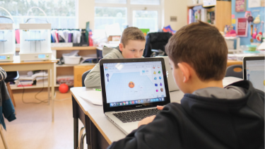 Two male students in a classroom studiously 3d modelling on Macbooks with 3D printers in the background.